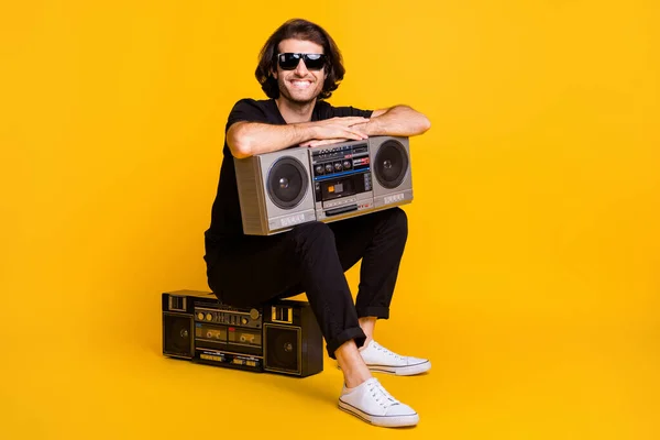 Foto de comprimento total de jovem segurar rádio vintage sentar boombox desgaste preto t-shirt calças tênis branco vidro solar isolado cor amarela fundo — Fotografia de Stock