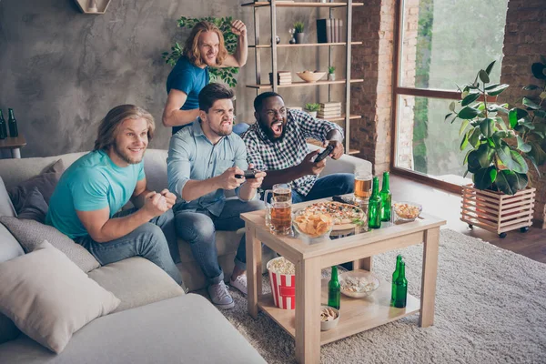 Foto de cuerpo entero de chico centrado sentarse acogedor sofá jugar joystick quieren ganar dos amigos pelo rubio apoyo levantar los puños tienen mesa con patatas fritas botella de cerveza en casa en el interior — Foto de Stock