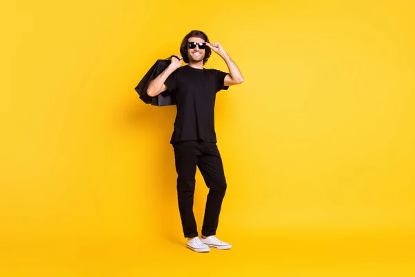 Фотографія молодого чоловіка, який тримає пакети для покупок, носить чорні футболки штани білі кросівки сонцезахисні окуляри ізольовані жовтого кольору — стокове фото