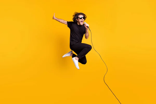 Foto de longitud completa de hombre joven salto levantar la mano micrófono boca abierta desgaste negro camiseta pantalones zapatillas blancas sunglass aislado color amarillo fondo — Foto de Stock