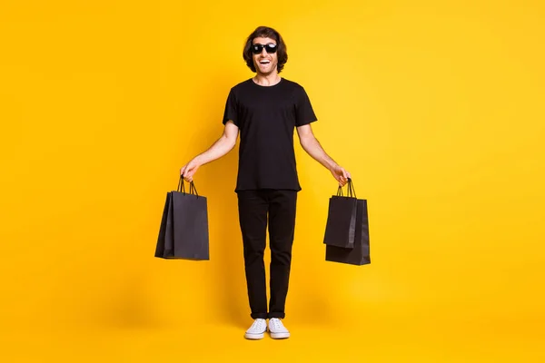 Ganzkörpergröße Foto von jungen Mann posiert halten Shop Pakete tragen schwarzes T-Shirt Hose weiße Turnschuhe Brille isoliert gelb Farbe Hintergrund — Stockfoto