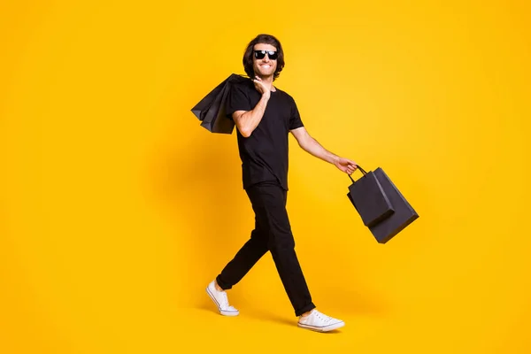 Volledige foto van jonge man lopen beer winkelcentrum tassen dragen zwarte t-shirt broek witte sneakers zonneglas geïsoleerde gele kleur achtergrond — Stockfoto