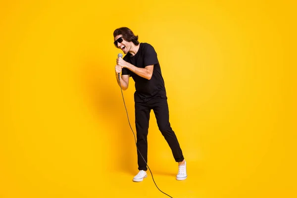 Full length foto di giovane uomo tenere mic cantare bocca aperta usura nero t-shirt pantaloni bianco scarpe da ginnastica occhiali isolato colore giallo sfondo — Foto Stock