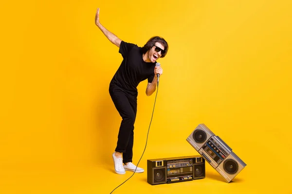 Фотографія молодої людини, що тримає мікрофон з відкритим ротом, піднімає руку два ретро радіоприймачі чорна футболка штани білі кросівки окуляри ізольовані жовтого кольору — стокове фото