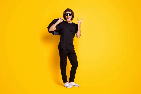 Ganzkörpergröße Foto von jungen Mann halten Pakete zeigen Hörner tragen schwarzes T-Shirt Hose weiße Turnschuhe Sonnenbrille isoliert gelbe Farbe Hintergrund — Stockfoto