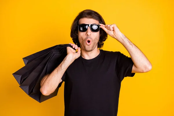 Foto di giovane uomo tenere negozio pacchetto shock espressione usura nero t-shirt sunglass isolato colore giallo sfondo — Foto Stock