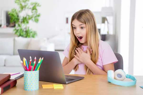 Foto de niña asombrada estudio uso remoto portátil impresionado profesor de escuela en casa noticias toque manos pecho sentarse mesa de confort escritorio en casa en interiores —  Fotos de Stock