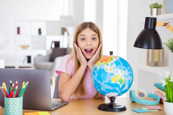 Photo of astonished kid girl study geography remote impressed new continent information scream touch hands face sit comfort table desk in house indoors