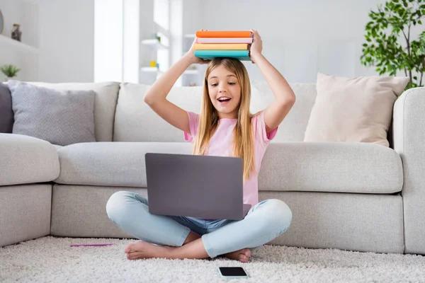 Foto de longitud completa de la computadora portátil remota de trabajo de niña pequeña emocionada tiene lección de tutor en línea reunión celebrar apilar libros cabeza sentarse piso piernas cruzadas dobladas en la casa en el interior — Foto de Stock