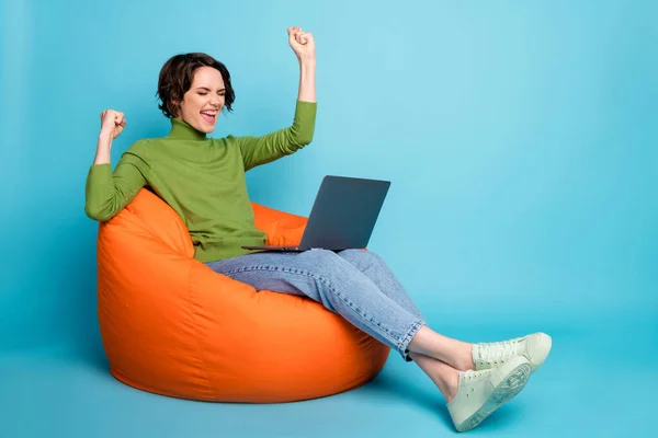 Full size profile side photo of delighted girl sit bean chair work laptop raise fsts wear autumn style pullover denim jeans isolated over blue color background — стоковое фото