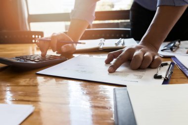 İş adamı rapor kağıdında işaret eder ve ofisteki ahşap masada analiz için hesap makinesi kullanır.