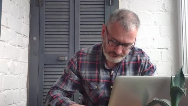 Gray-haired bearded male accountant working at home behind a laptop, making a report against the backdrop of a minimalist interior. Verifies data with the monitor and writes in a notebook. — Stock Video