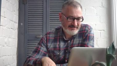 Gri saçlı sakallı erkek muhasebeci bir dizüstü bilgisayar, evde çalışan bir rapor bir minimalist zemin karşı iç yapım, Skype, içecek çay veya kahve, gülümser bir müşteriden bir çağrı alır ve konuşuyor.