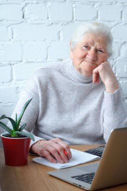 Yaşlı bir kadın laptop önünde masada oturuyor.