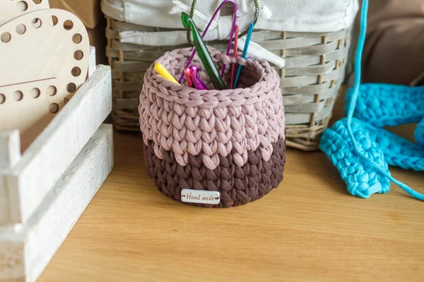 Two-tone knitted basket on a light wooden surface against the background of knitting and details for making handmade knitted bags. — Stock Photo, Image