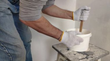 Gri saçlı sakallı bir adam bir kova bir sopa ile beyaz boya karışımları ve konteyner içine, bir odada duvar boyama için hazırlama.