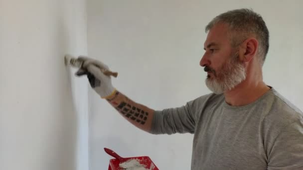 髭の男は ペイントブラシを使用してフラットで内壁を塗装します 修理をしてハンサムな若い男 積極的に白いペンキで壁を塗ります ホームリノベーションまたは模様替えコンセプト — ストック動画