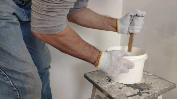 白髪のひげを生やした男が バケツの中の白いペンキを棒で混ぜ合わせ 容器に注ぎ 部屋の壁を塗るための準備をする — ストック動画