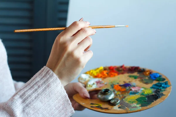 Close-up of female hands in a pink sweater holding a brush and a palette with multi-colored acrylic paints against a white canvas — Stock Photo, Image