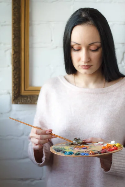 Young woman with long black hair holds a brush and a palette with multi-colored acrylic paints on the background of an empty vintage frame — Stock Photo, Image