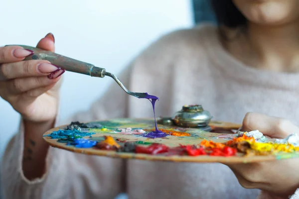 Close-up of female hands mixing paints on a palette with a spatula against a white canvas, creating an oil painting — Stock Photo, Image