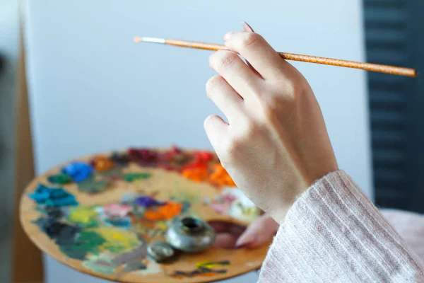 Close-up of female hands in a pink sweater holding a brush and a palette with multi-colored acrylic paints against a white canvas — Stock Photo, Image