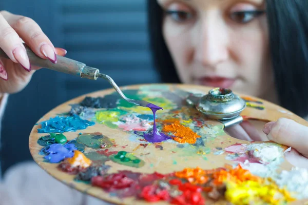 Closeup of a young woman with black hair mixes paint on a palette with a spatula against a white canvas, creating an oil painting — Stock Photo, Image