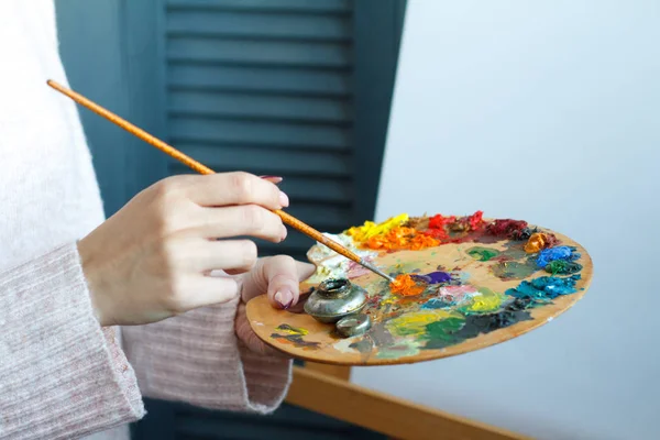 Close-up of female hands in a pink sweater holding a brush and a palette with multi-colored acrylic paints against a white canvas — Stock Photo, Image
