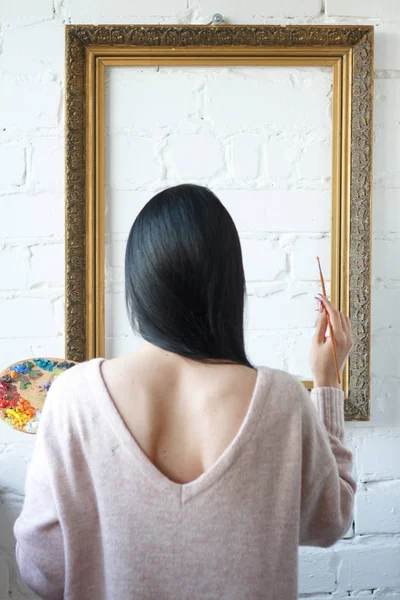 A young woman with long black hair is holding her back holding a brush and a palette with colorful paints on a background of an empty vintage frame — Stock Photo, Image