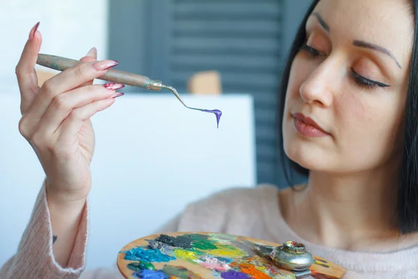 Closeup of a young woman with black hair mixes paint on a palette with a spatula against a white canvas, creating an oil painting — Stock Photo, Image