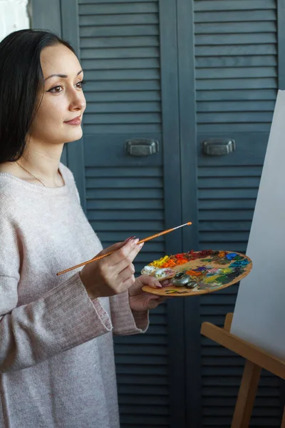 Eine junge Frau östlichen Aussehens mit langen schwarzen Haaren hält einen Pinsel und eine Palette mit mehrfarbigen Acrylfarben in den Händen — Stockfoto