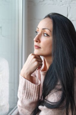 Closeup uzun siyah saçlı gülümseyen güzel genç kadın pencereden dışarı bakar, pencereye yansıyan