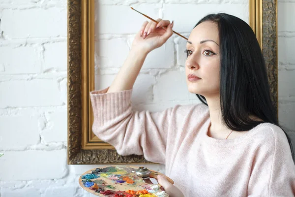 Young woman with long black hair holds a brush and a palette with multi-colored acrylic paints on the background of an empty vintage frame — Stock Photo, Image