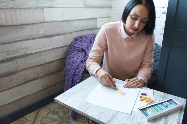 A girl with long black hair in a pink sweater draws with multicolored crayons on a white sheet — Stock Photo, Image