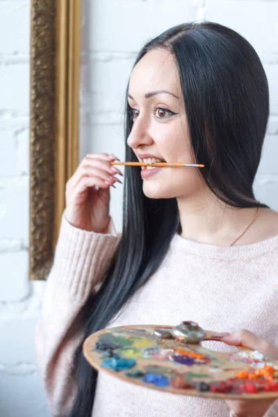 Крупним планом молода жінка з чорними сумішами волосся малює на палітрі пензлем на тлі порожньої старовинної рамки, створюючи олійну картину — стокове фото