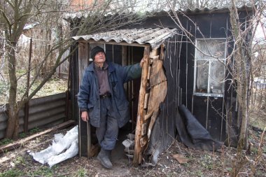 Özensiz giysiler içinde yaşlı bir adam kendi harap evinin eşiğinde duruyor ve mesafe içine bakar