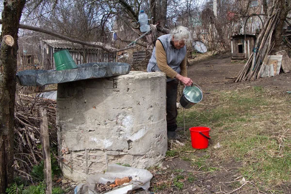 Una anciana en un pueblo desierto está recogiendo agua de un pozo en un cubo, viviendo sola —  Fotos de Stock