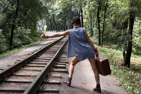 Young girl with pigtails in a blue shirt with a suitcase walks on the rails of the railway — Stock Photo, Image