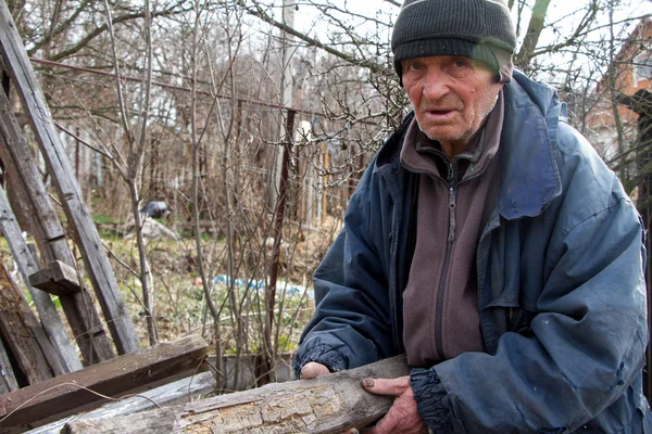 Un anciano con ropa desordenada elige entre una pila de tablas de madera viejas para calentar la casa, la soledad en el pueblo —  Fotos de Stock
