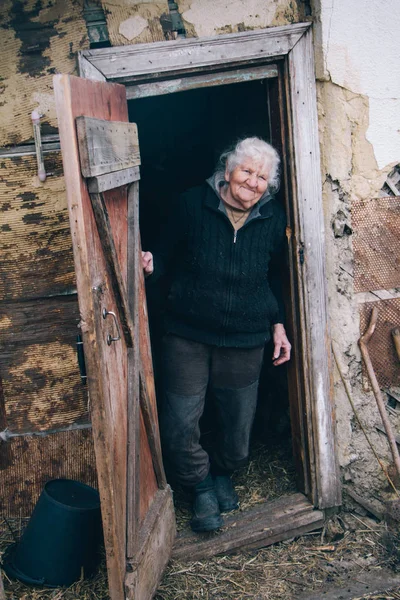 Дуже стара жінка з сірим волоссям у старому одязі стоїть на порозі старенького будинку — стокове фото