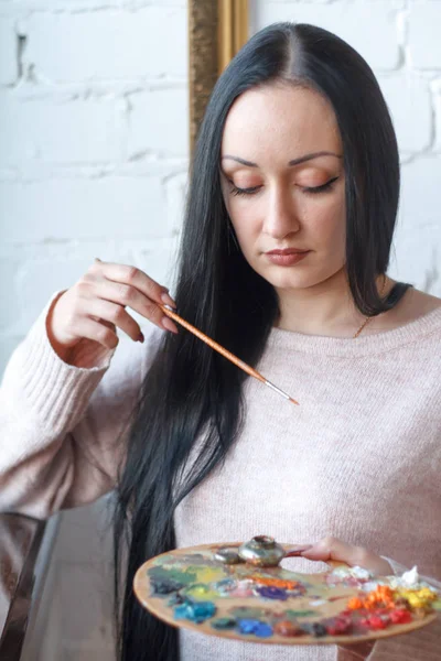 Closeup of a young woman with black hair mixes paint on a palette with a brush against a background of an empty vintage frame, creating an oil painting — Stock Photo, Image