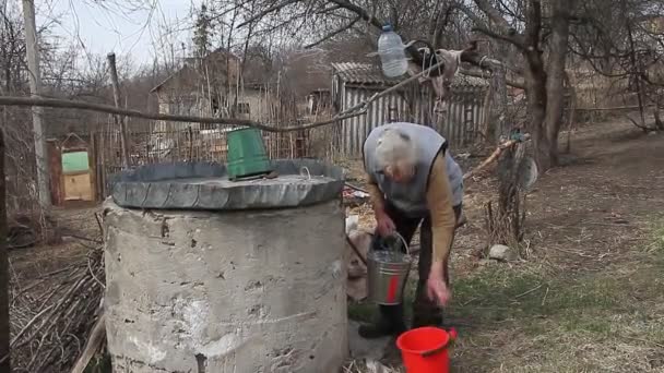 En gammal kvinna i en öde by samlar vatten från en brunn i en hink, bor ensam — Stockvideo