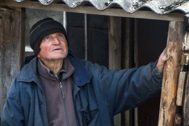Özensiz giysiler içinde yaşlı bir adam kendi harap evinin eşiğinde duruyor ve mesafe içine bakar