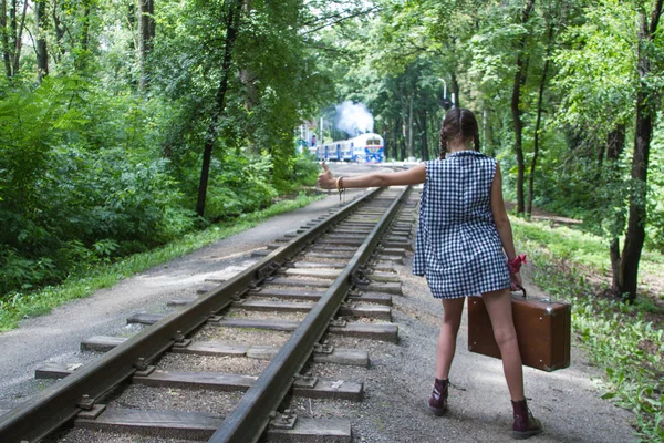 Young girl with pigtails in a blue shirt with a suitcase walks on the rails of the railway — Stock Photo, Image