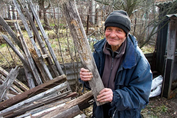 Un anciano con ropa desordenada elige entre una pila de tablas de madera viejas para calentar la casa, la soledad en el pueblo —  Fotos de Stock