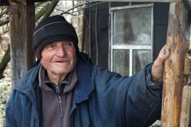 Özensiz giysiler içinde yaşlı bir adam kendi harap evinin eşiğinde duruyor ve mesafe içine bakar