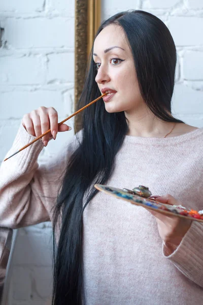 Closeup of a young woman with black hair mixes paint on a palette with a brush against a background of an empty vintage frame, creating an oil painting — Stock Photo, Image