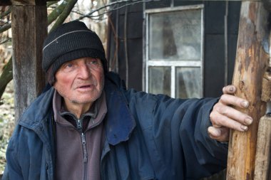 Özensiz giysiler içinde yaşlı bir adam kendi harap evinin eşiğinde duruyor ve mesafe içine bakar
