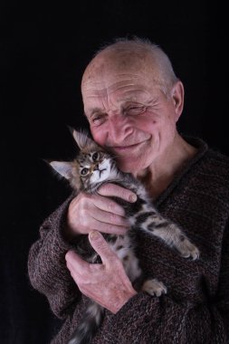 Yaşlı bir adam yüzüne Maine Coon kedi yavrusu tutar ve gülümsüyor, siyah bir arka plan, seçici odak.