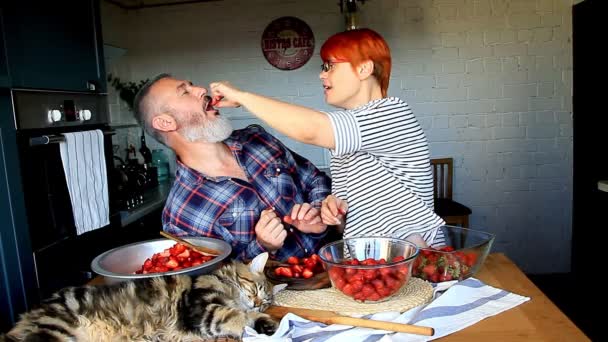 成年夫妇男人和女人剥草莓果酱和切草莓果酱 互相喂食 笑和有乐趣 缅因库恩小猫睡在厨房的桌子上 — 图库视频影像